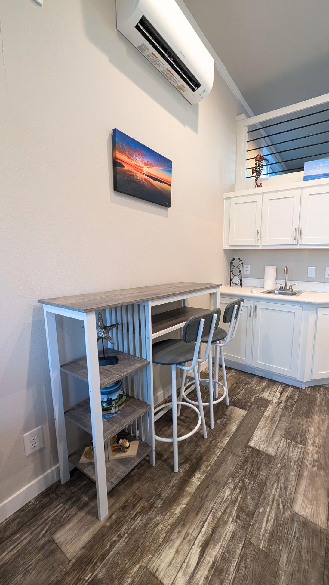 Small dining or workspace area with a narrow table, two stools, shelving, and white kitchen cabinets nearby under a wall-mounted air conditioning unit.