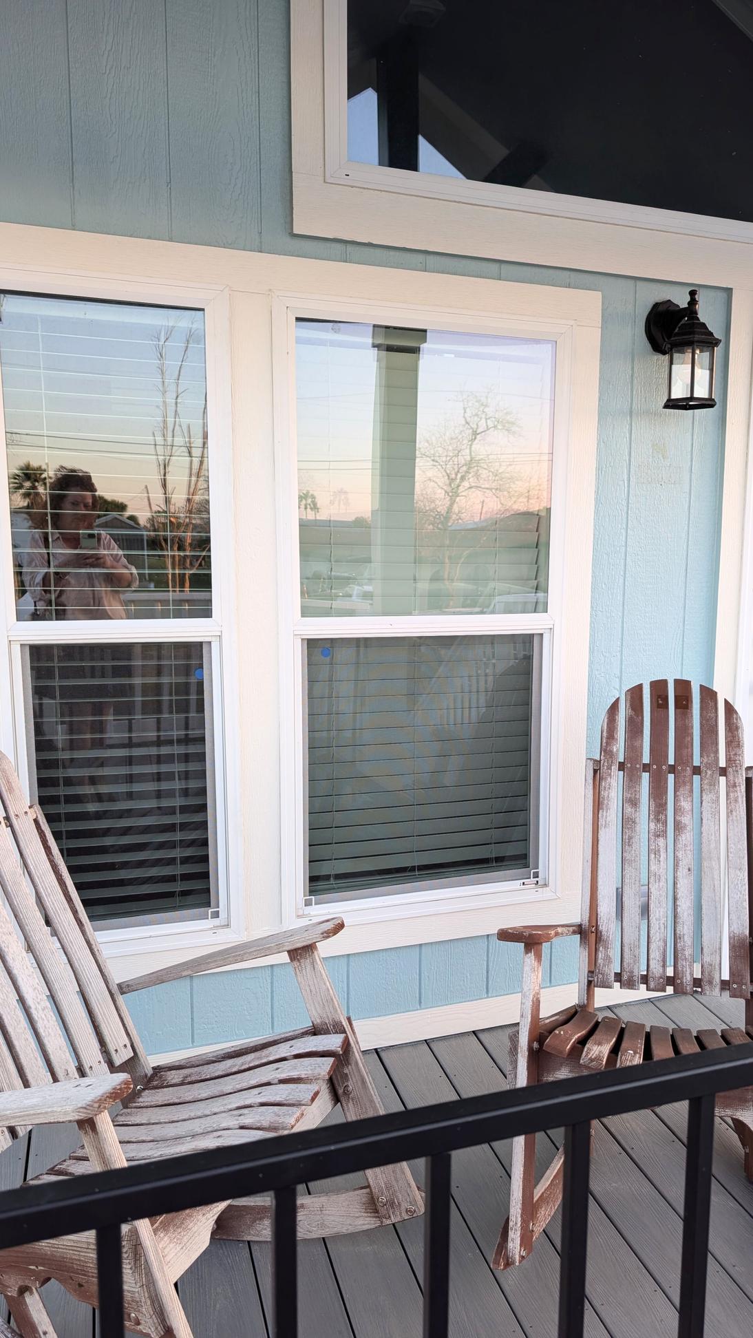 Two wooden rocking chairs on a porch in front of large windows with white trim, light blue siding, and an outdoor wall lantern.