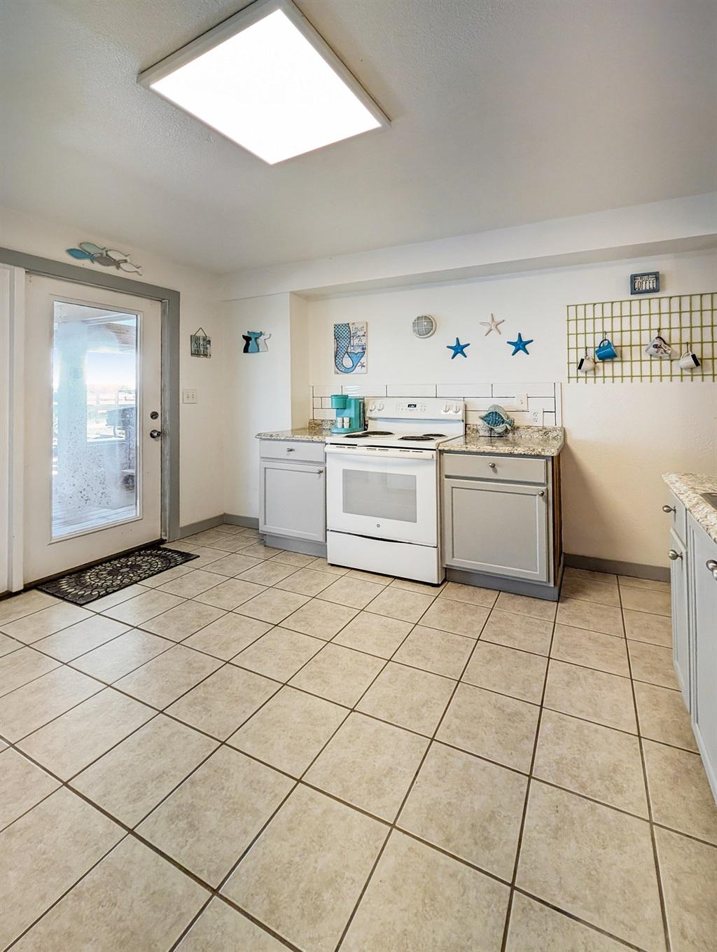 Bright kitchen with white stove, gray cabinets, tile flooring, coastal wall decor, and a glass door leading to an outdoor area.