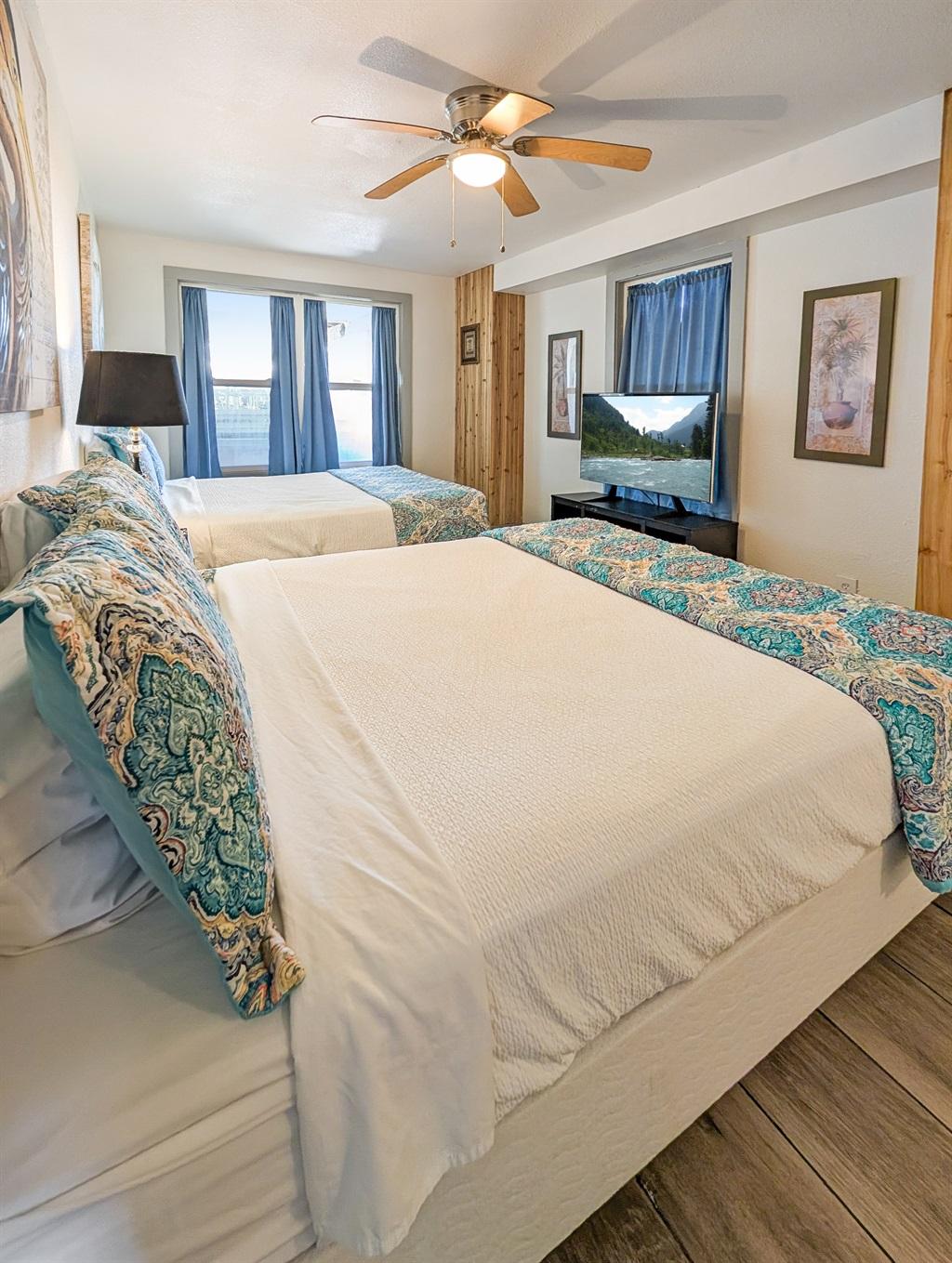 Bedroom with two large beds, patterned pillows, a TV on a stand, ceiling fan, and windows with curtains overlooking the water.