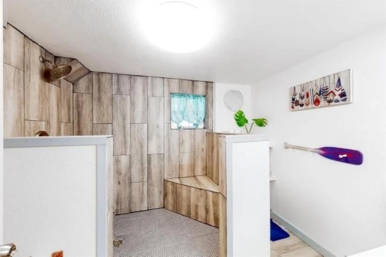 Large walk-in shower with wood-look tile walls, built-in bench, small window with curtain, and decorative coastal accents.