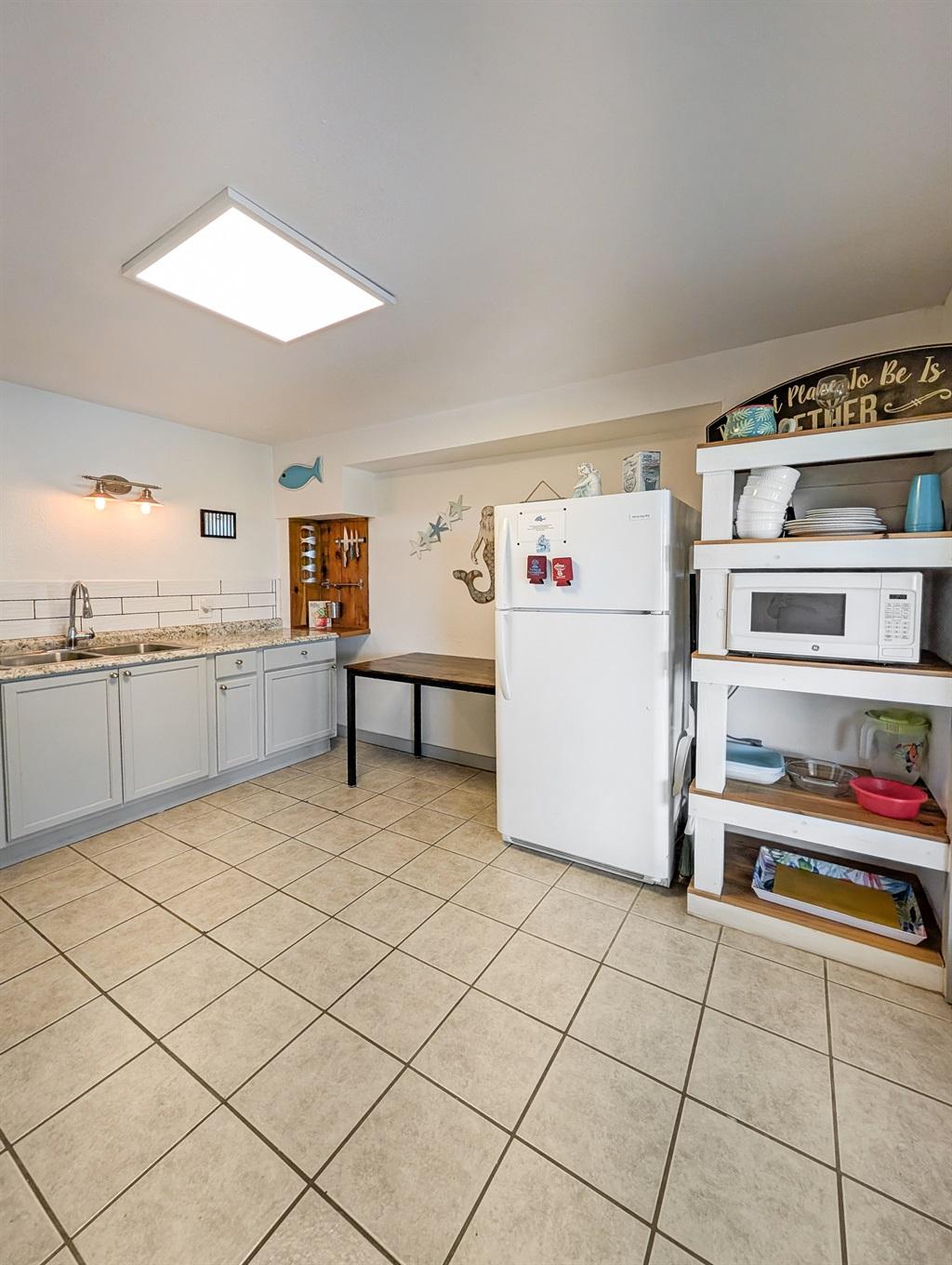 Large kitchen with gray cabinets, white refrigerator, open shelving with microwave and dishes, tile flooring, and coastal decor accents.
