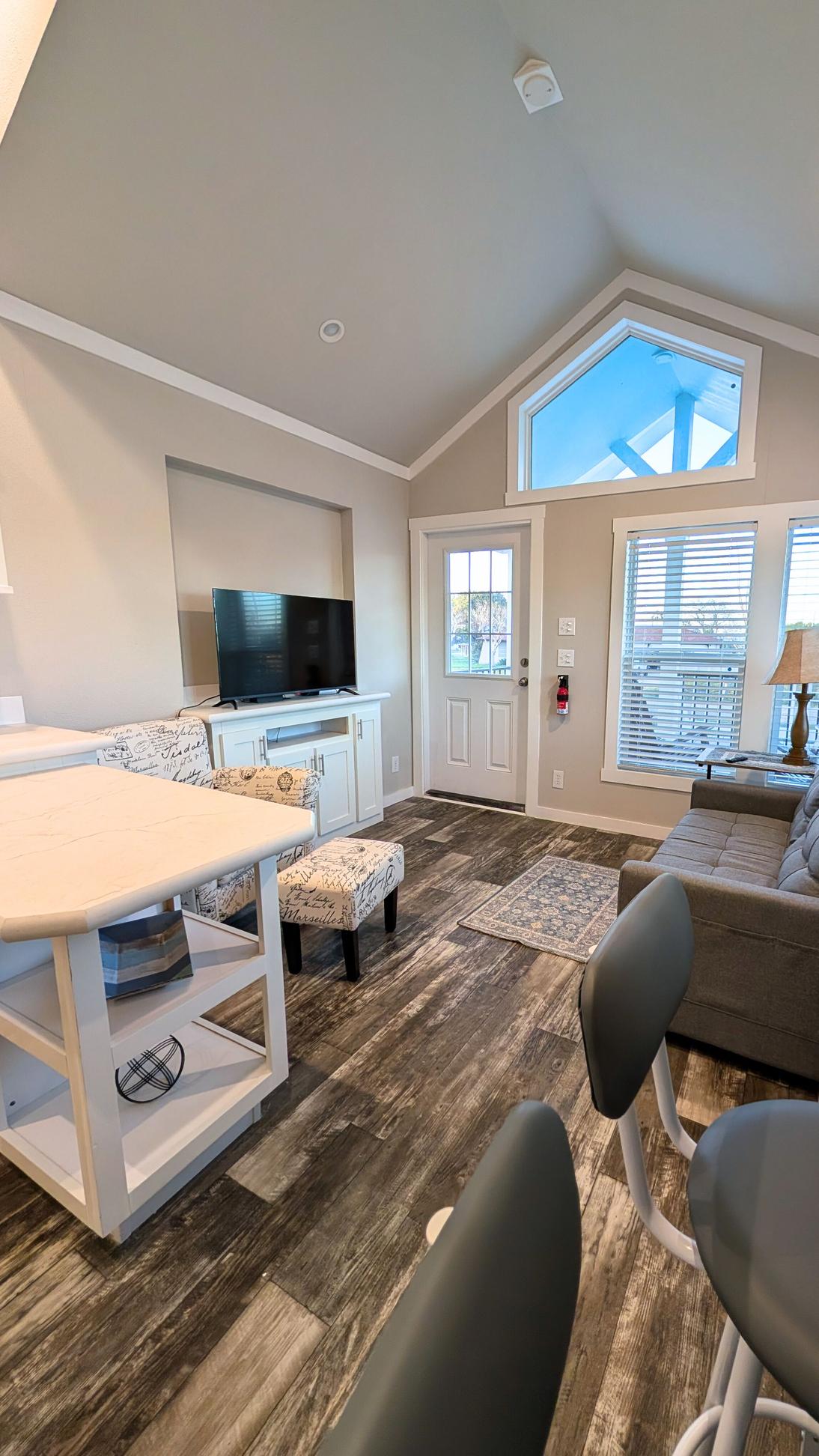 Bright open living area with vaulted ceiling, large windows, TV on built-in cabinet, kitchen island with seating, and a sofa near the front door.