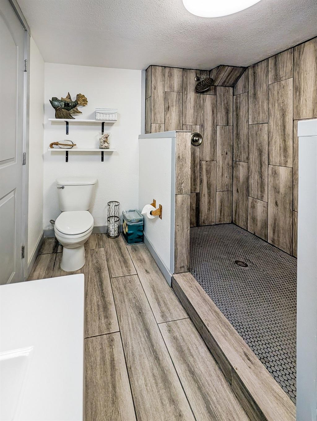 Bathroom with a large walk-in shower featuring wood-look tile walls and pebble-style floor, toilet with shelving above, and matching tile flooring throughout.