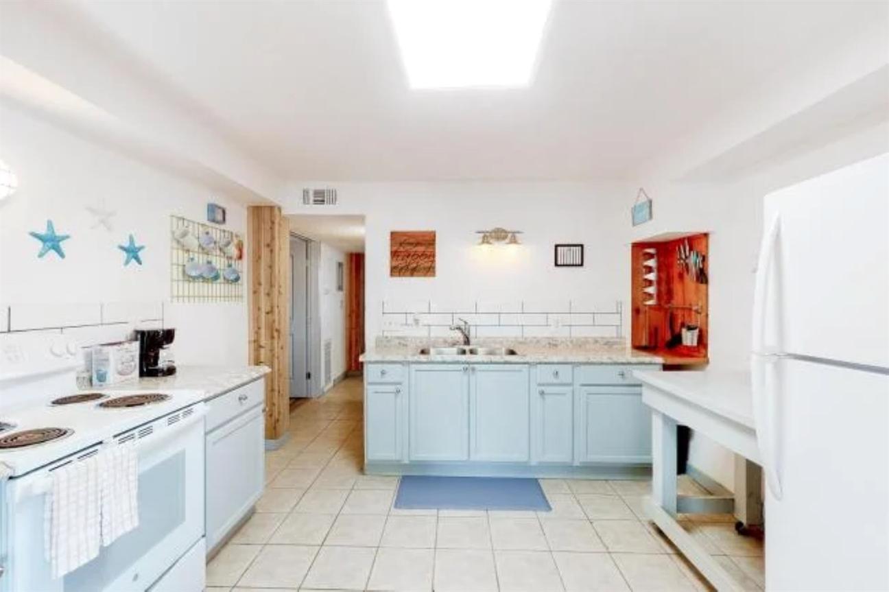 Wide kitchen with white appliances, light blue cabinets, long countertops, tile flooring, and coastal wall decor accents.