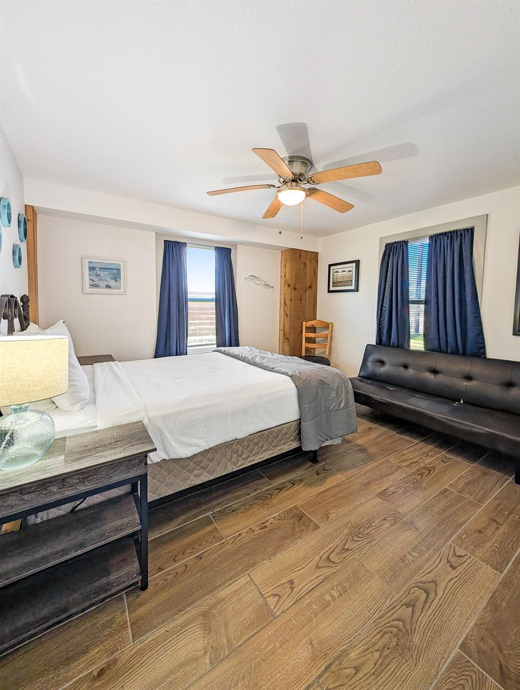 Bedroom with a bed, nightstand and lamp, black futon, wood-look flooring, ceiling fan, and windows with curtains offering views outside.