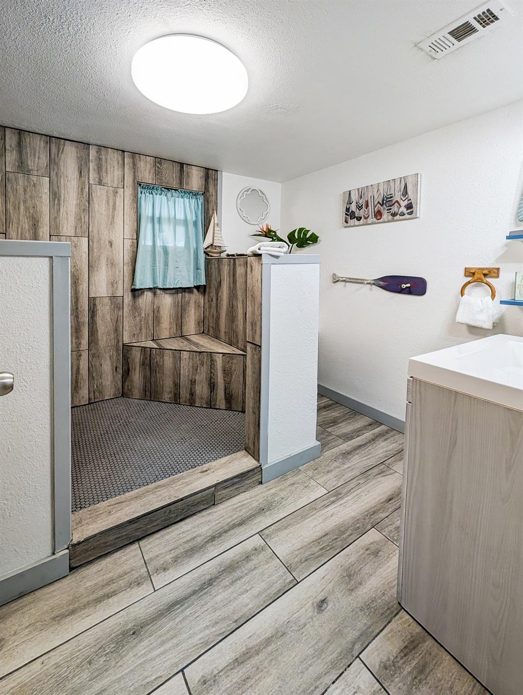 Bathroom featuring a large walk-in shower with wood-look tile, built-in bench, small window with curtain, vanity area, and wood-look flooring.
