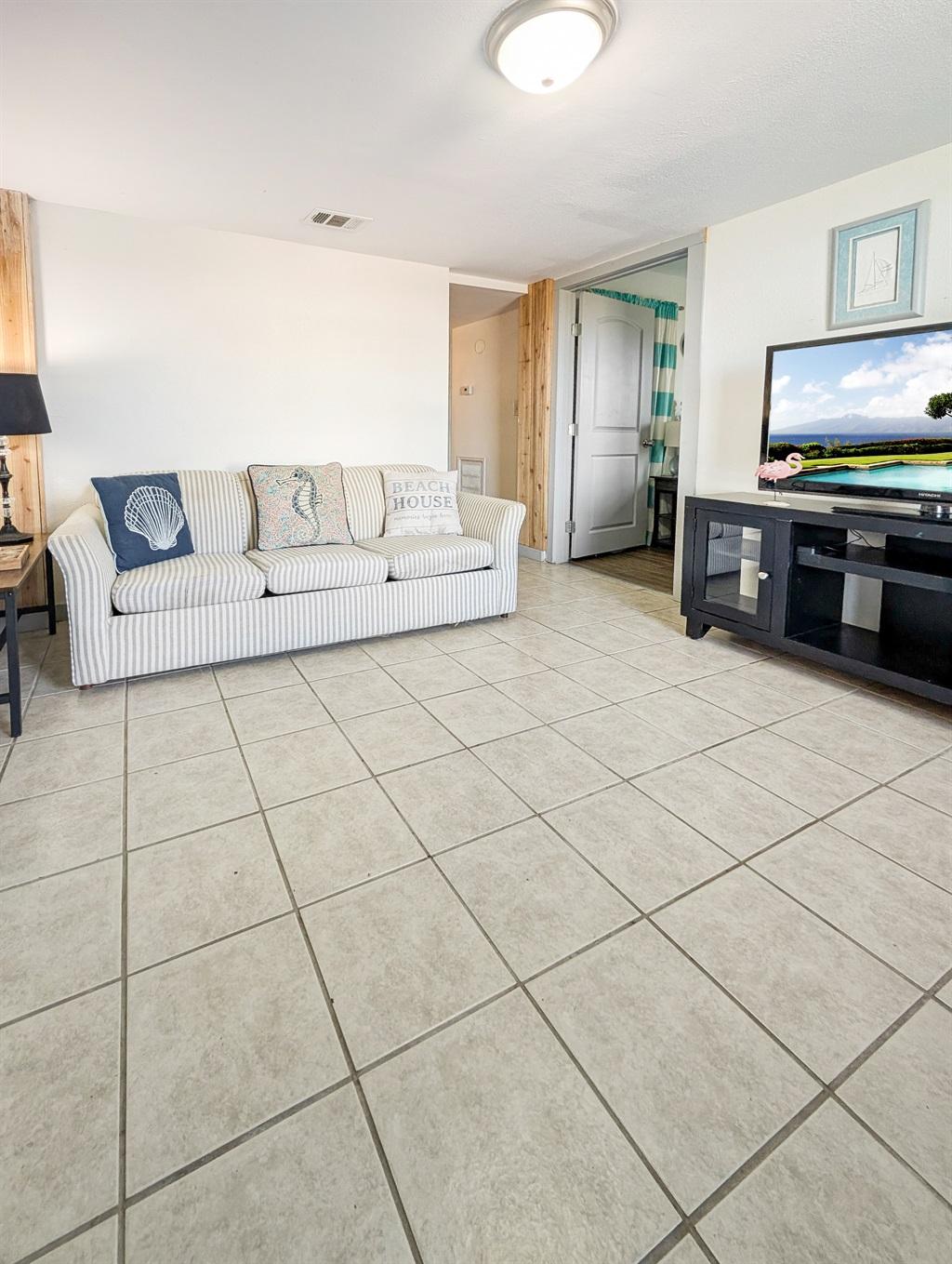 Living room with a striped sofa, decorative pillows, tile flooring, TV on a stand, and an open doorway leading to another room.