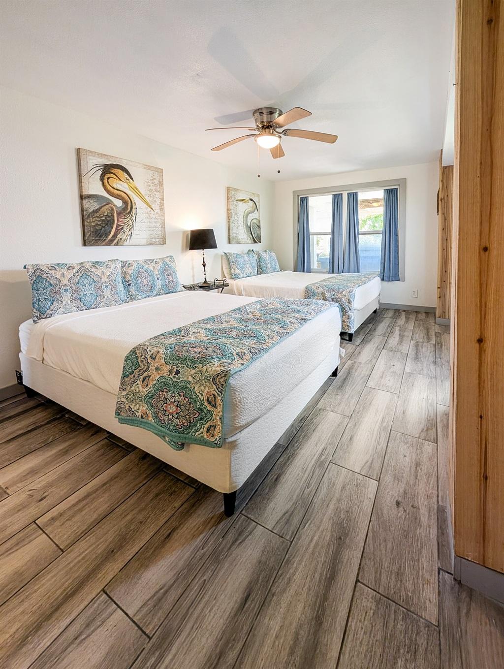 Bedroom with two beds featuring patterned bedding, wood-look flooring, wall art, a ceiling fan, and a window with curtains letting in natural light.