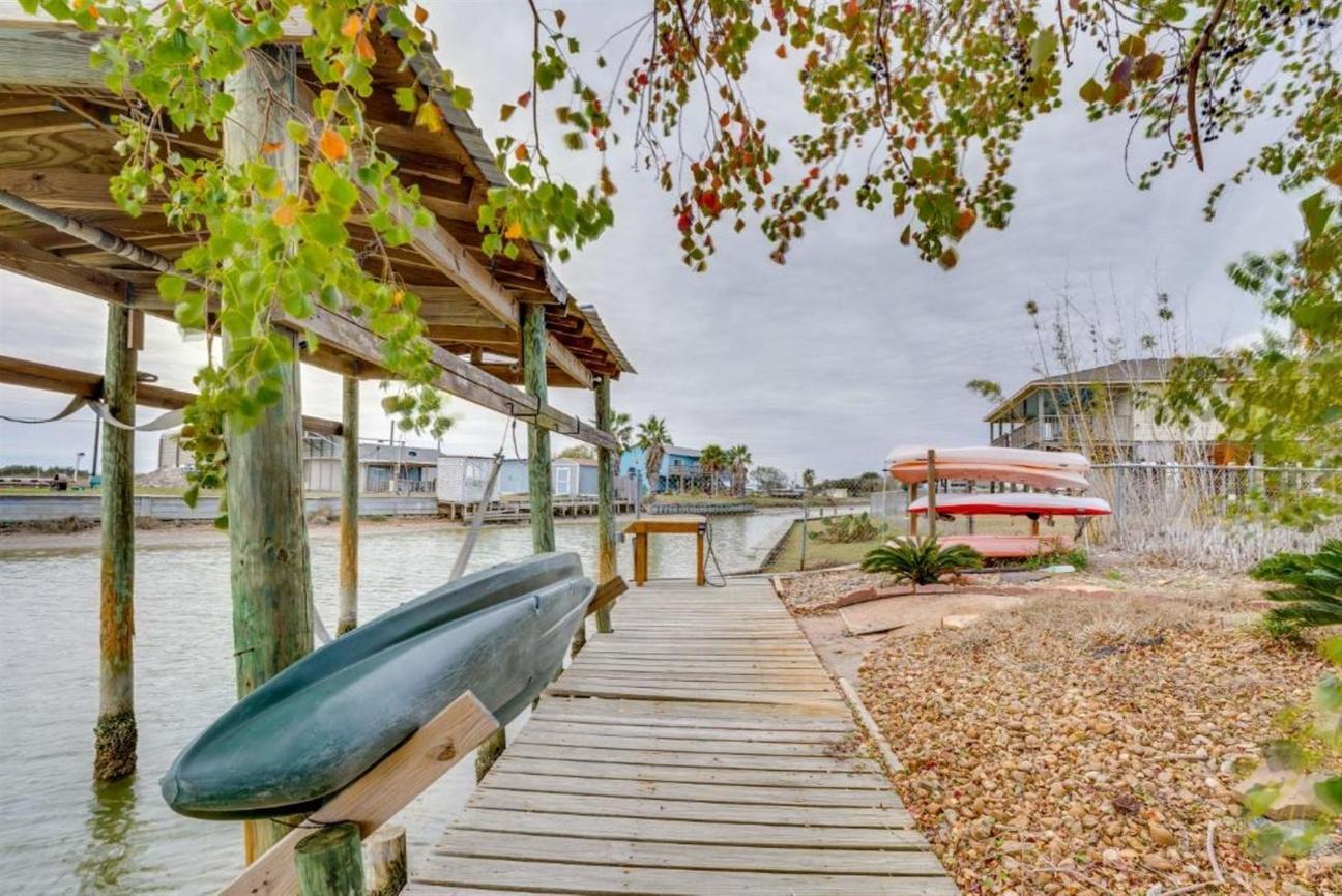 Wooden dock along the water with a kayak mounted on the side, nearby kayaks stacked onshore, and coastal homes in the background.