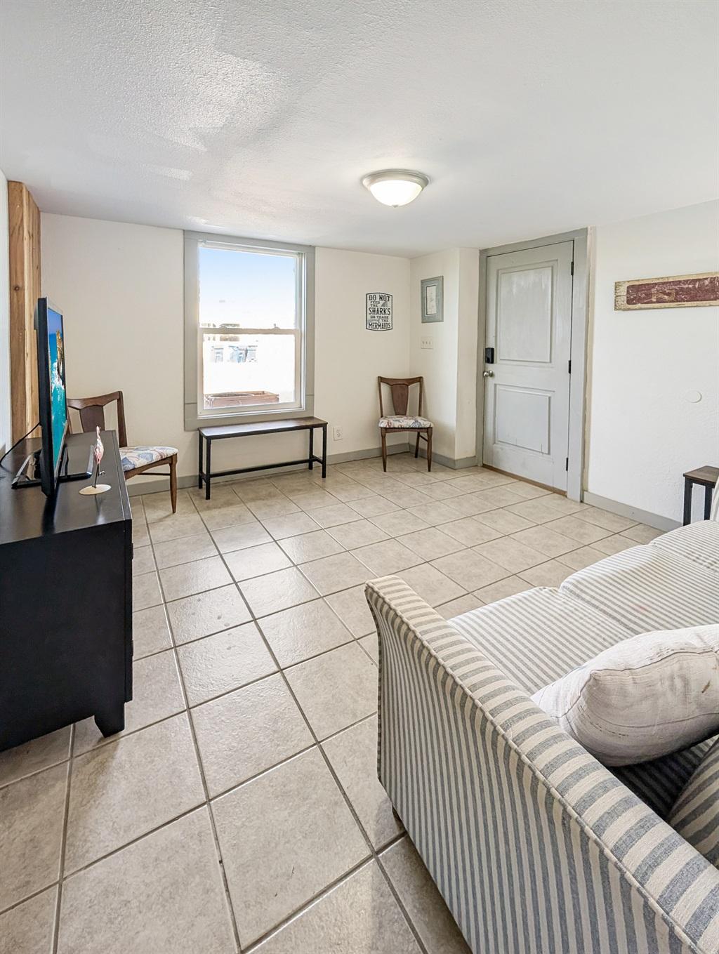 Living room with tile flooring, a striped sofa, TV on a stand, chairs near a window, and a door leading outside.