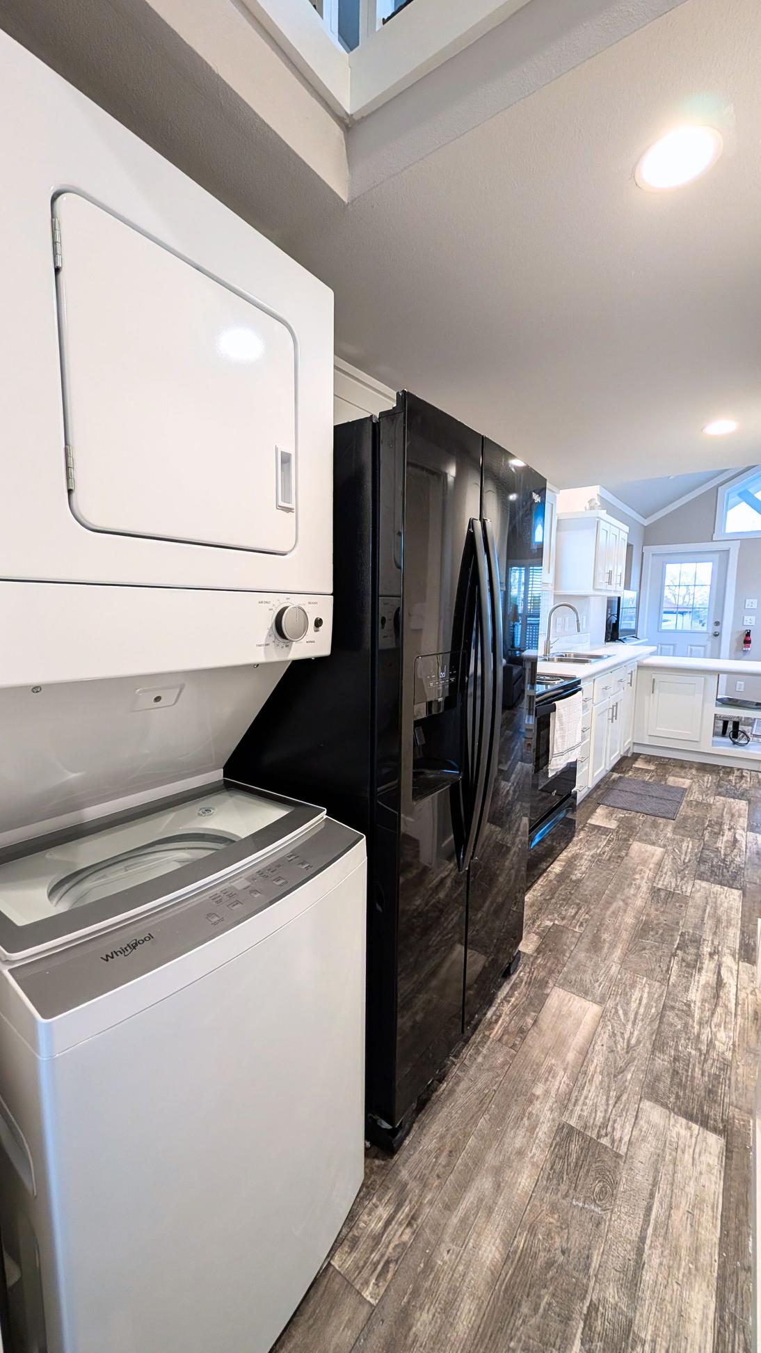 Stacked washer and dryer unit next to a black refrigerator in a modern kitchen with white cabinets and wood-look flooring.