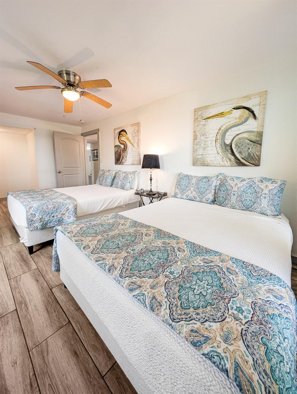 Bedroom with two large beds featuring patterned blue bedding, wood-look flooring, wall art of coastal birds, a ceiling fan, and a bedside table with a lamp.