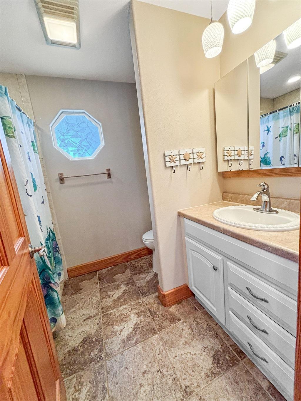 Bathroom with a white vanity and sink, large mirror with pendant lighting, shower curtain with coastal design, and tile flooring with an octagonal window.