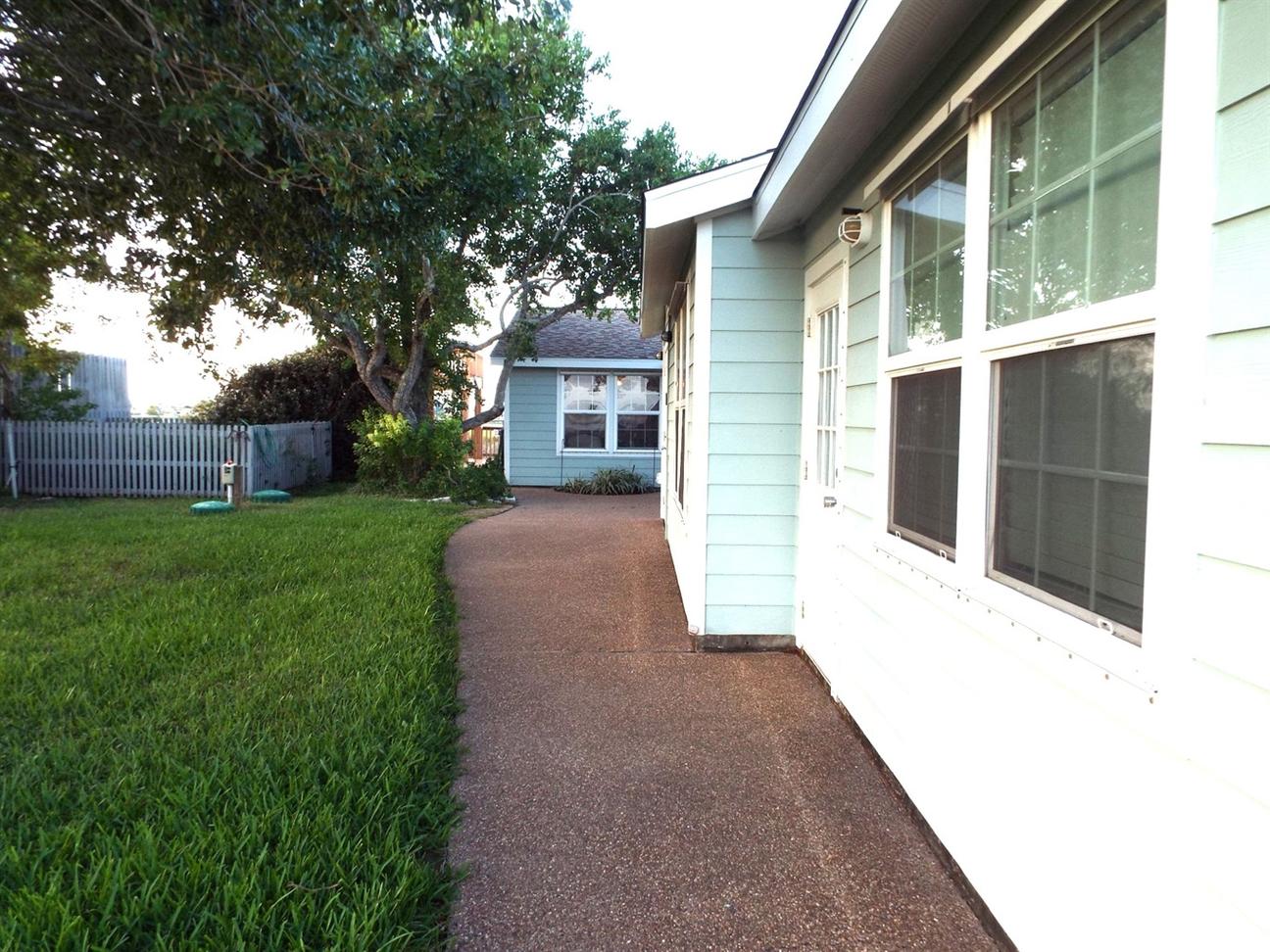 Side yard of a coastal home with a concrete walkway, green grass, trees for shade, and light blue exterior siding.
