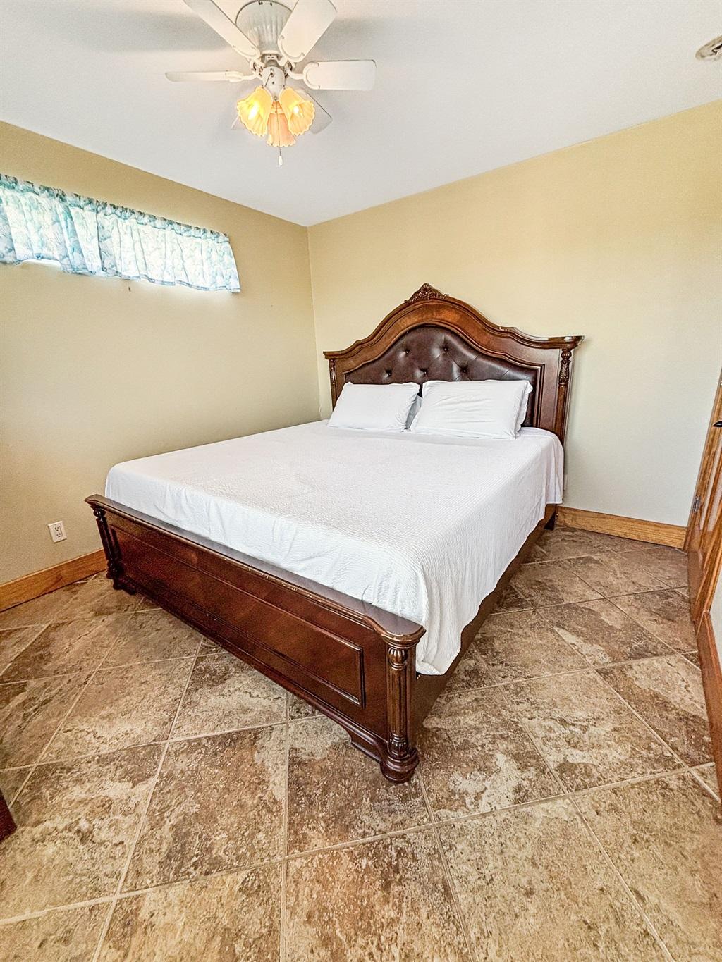 Bedroom with a wooden bed frame, white bedding, tile flooring, ceiling fan, and light-colored walls with a small window.