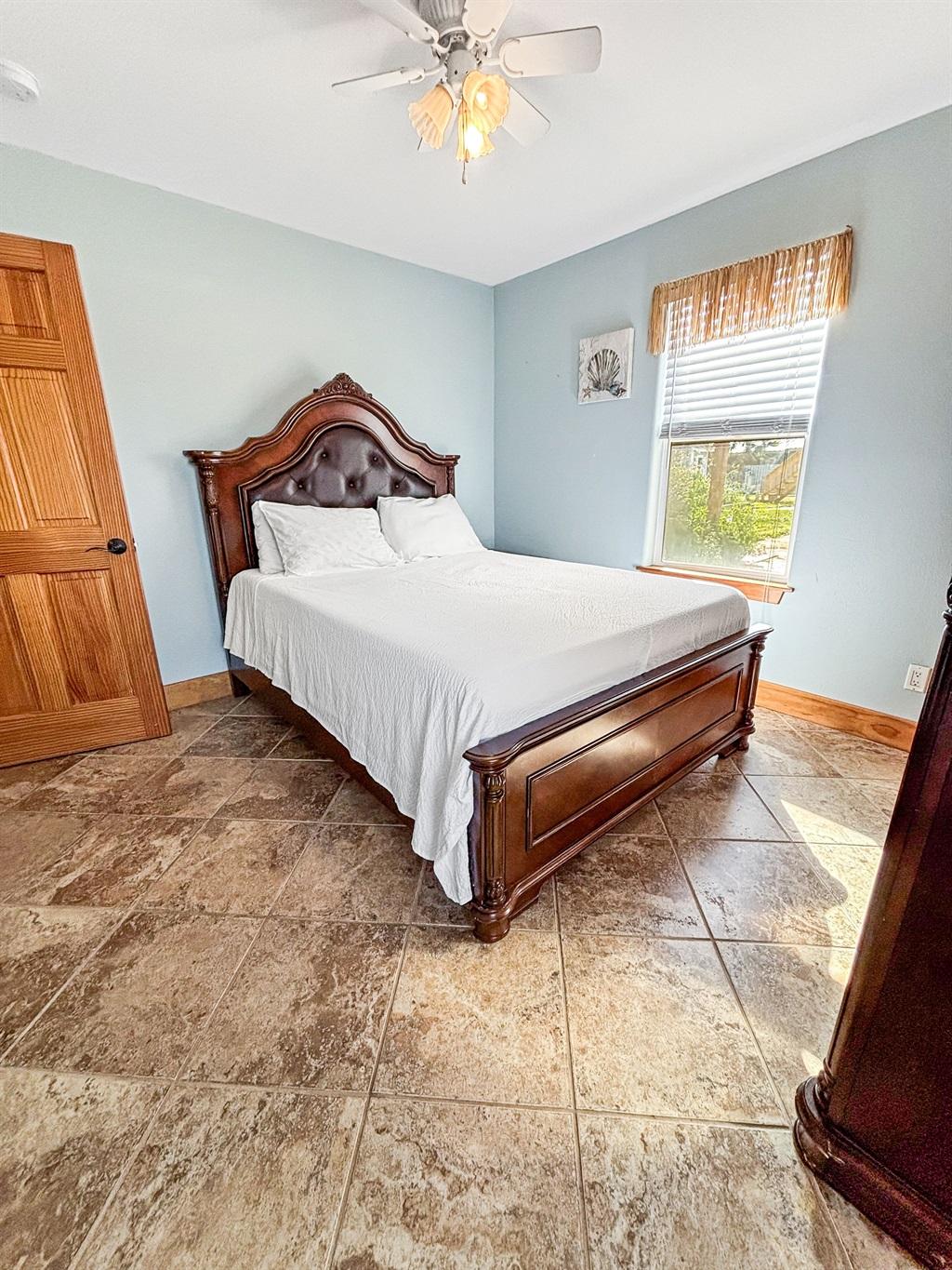 Bedroom with a wooden bed frame, white bedding, light blue walls, tile flooring, a window with natural light, and a ceiling fan overhead.