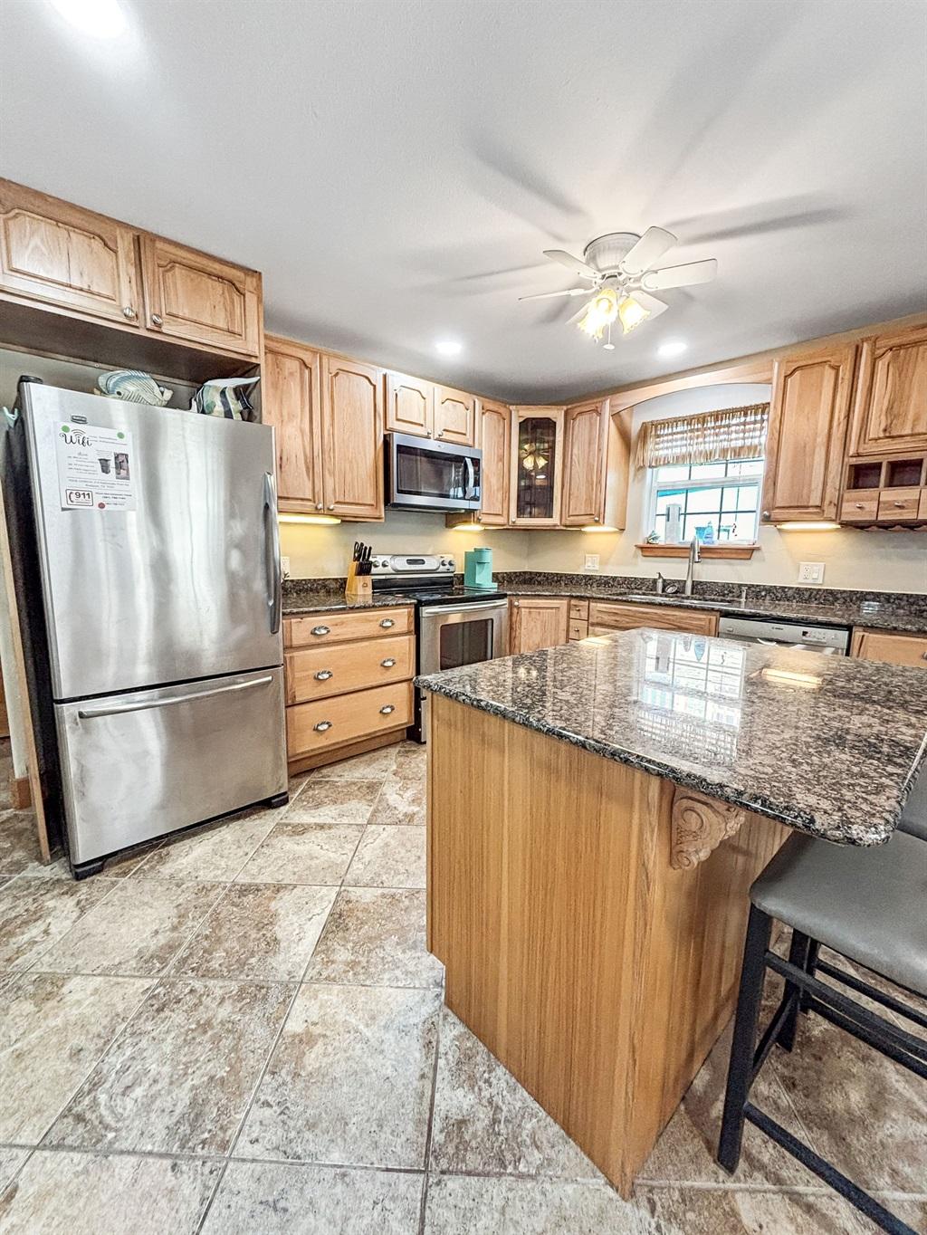 Large kitchen with wood cabinets, granite countertops, center island with seating, stainless steel refrigerator, and tile flooring.