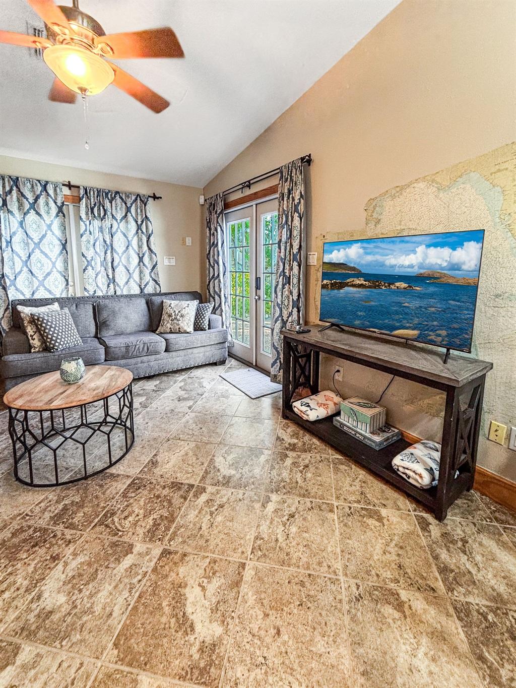 Living room with a gray sofa, round coffee table, TV on a console, ceiling fan, and glass doors leading outside, with tile flooring and natural light.