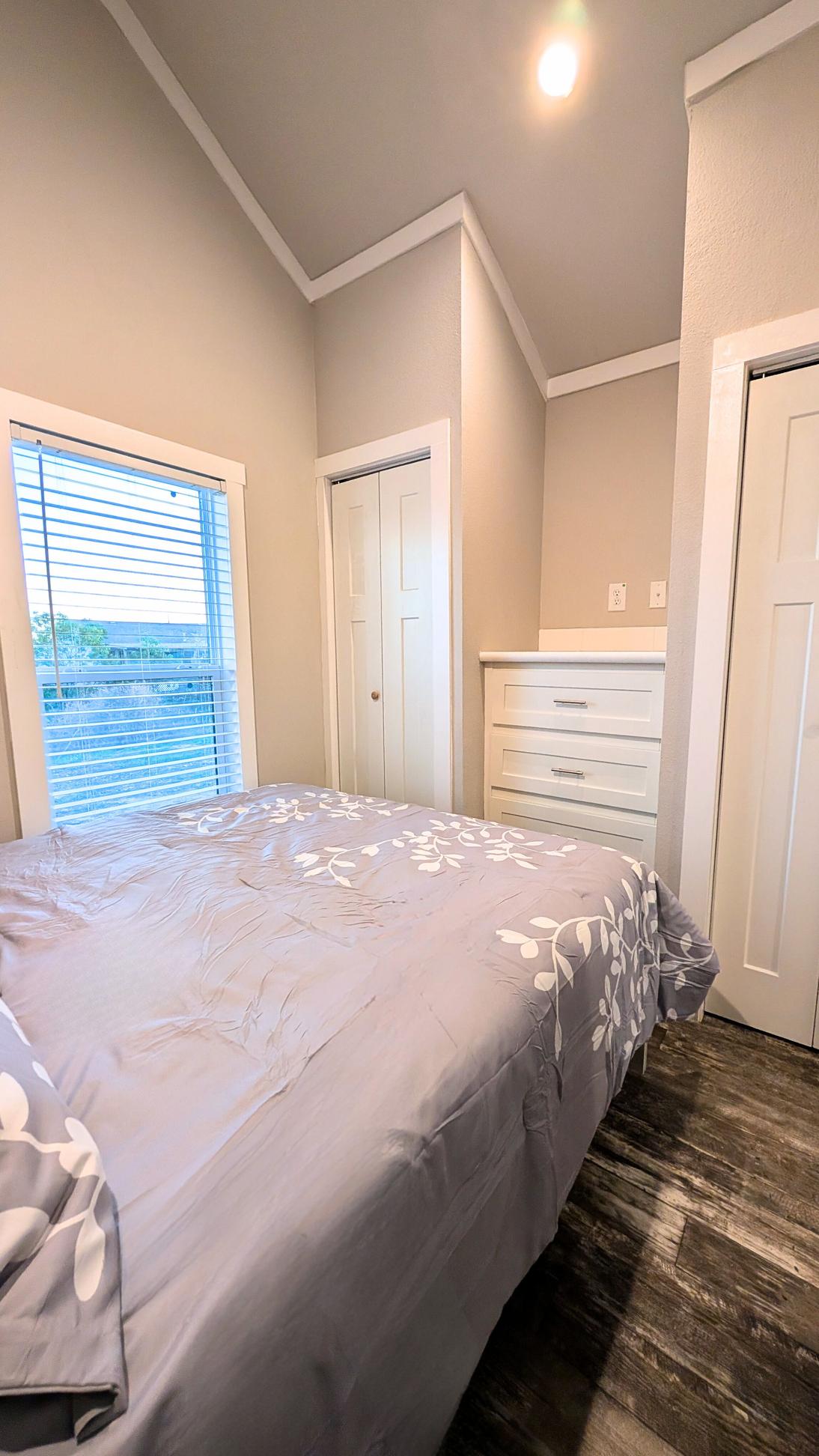 Bedroom with a bed featuring a gray patterned comforter, a window with blinds, and built-in drawers and closet doors in a neutral-toned space.