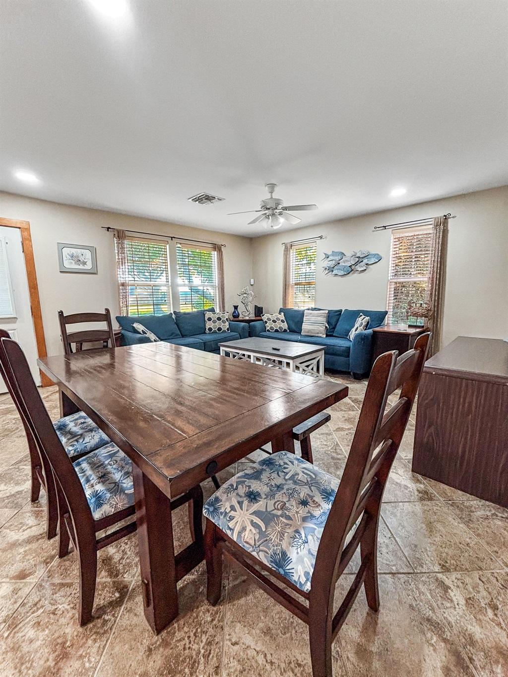 Dining area with a wooden table and chairs leading into a living room with a blue sectional sofa, coffee table, and multiple windows with natural light.