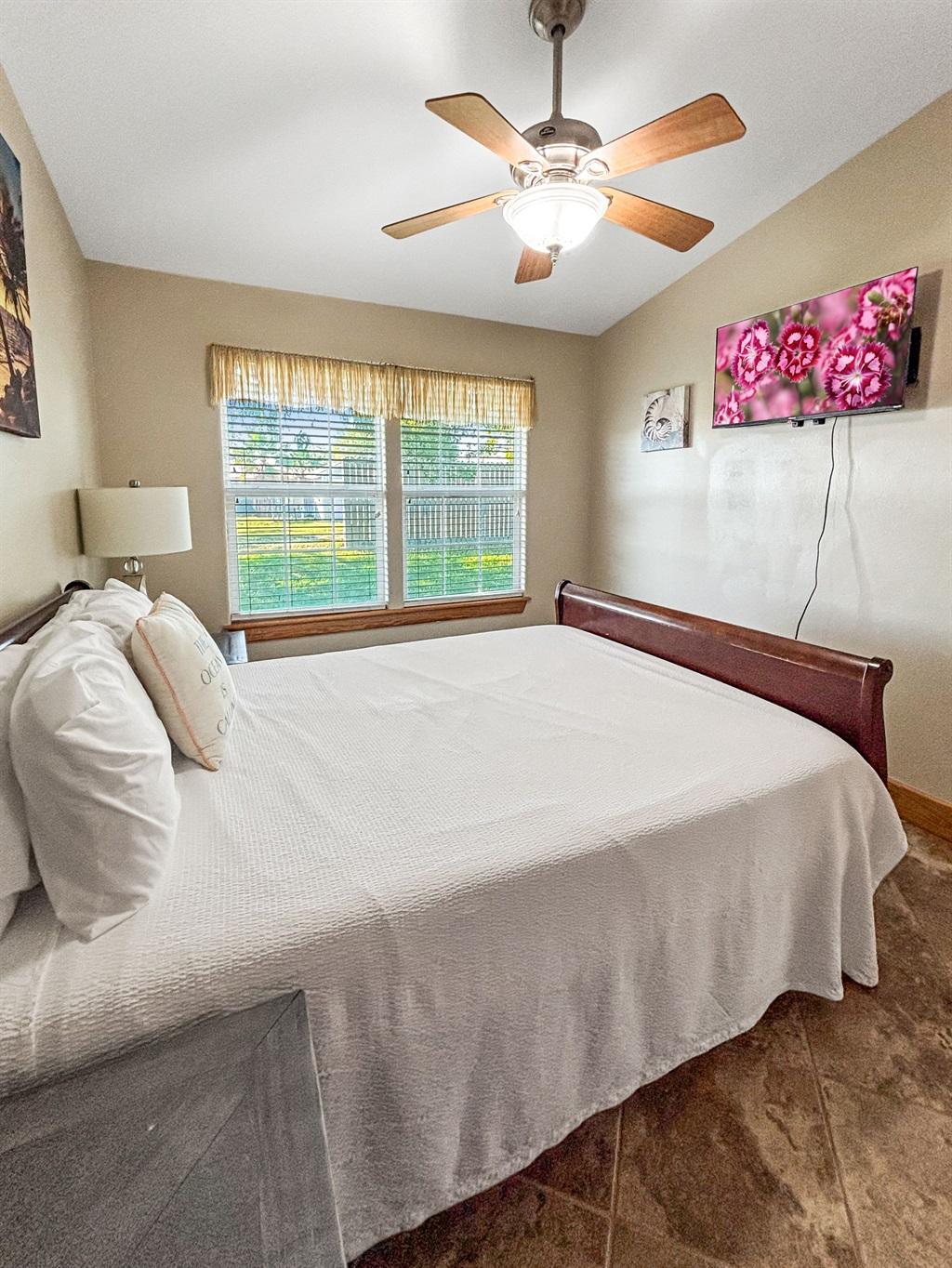 Bedroom with a bed featuring white bedding, a window with blinds, ceiling fan, bedside lamp, and a wall-mounted TV displaying flowers.