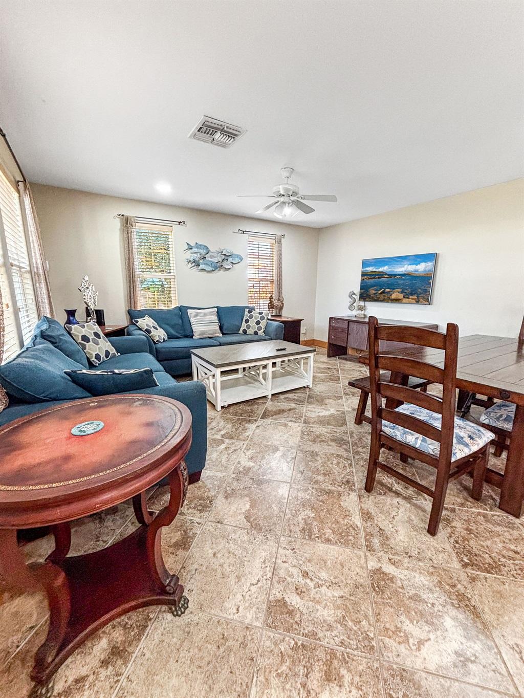 Living room with a blue sectional sofa, coffee table, side table, and tile flooring, adjacent to a dining area with wooden chairs and coastal wall decor.