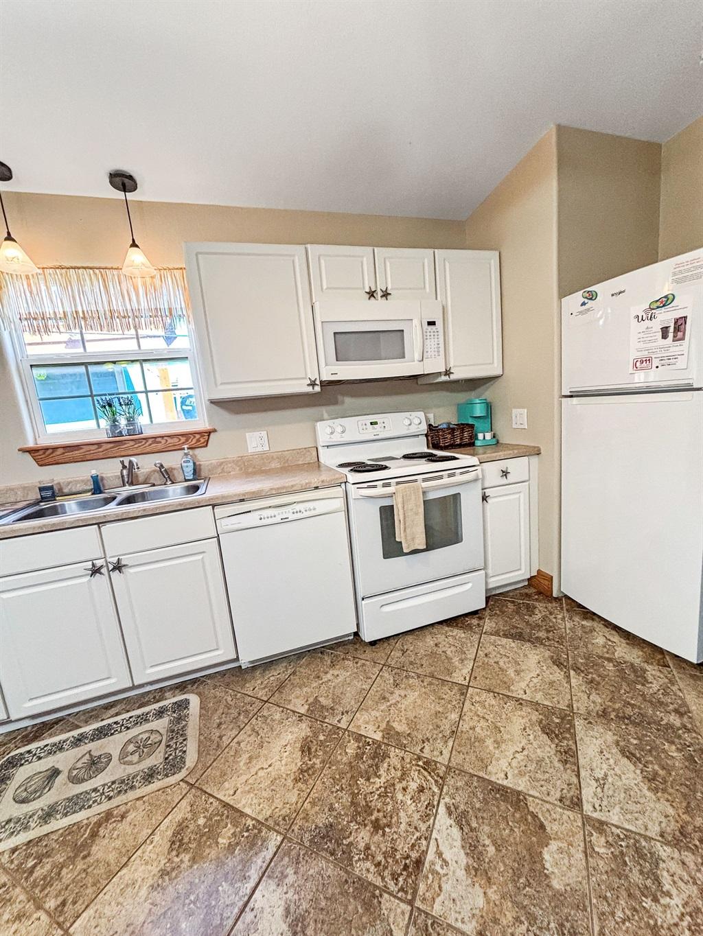 Kitchen with white cabinets, white stove and microwave, refrigerator, sink under a window, and tile flooring.