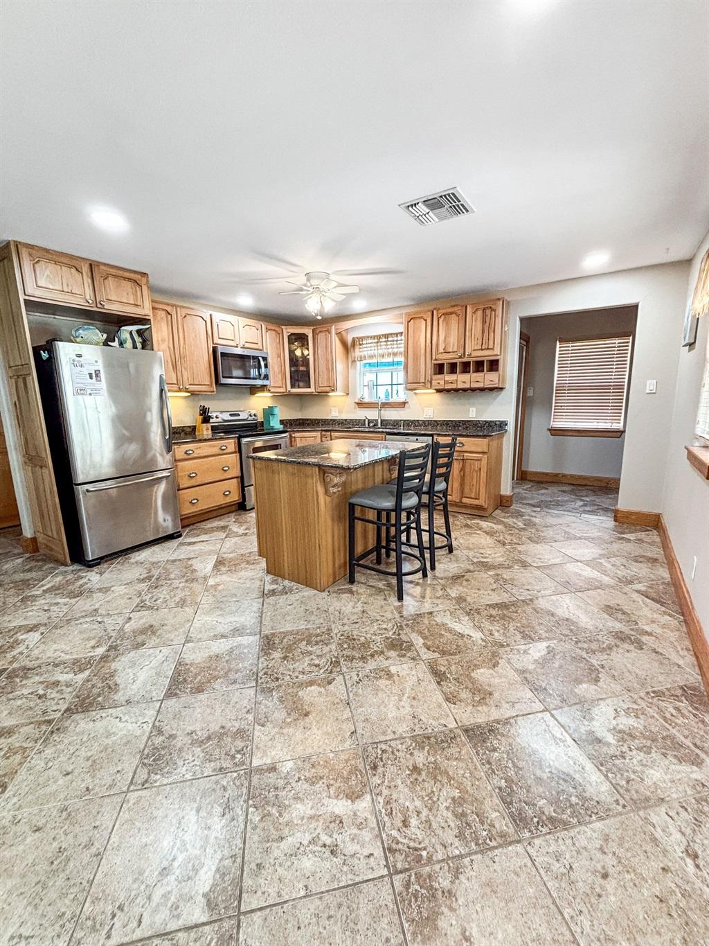 Large kitchen with wood cabinetry, granite island with bar stools, stainless steel refrigerator, tile flooring, and an open layout with ample space.
