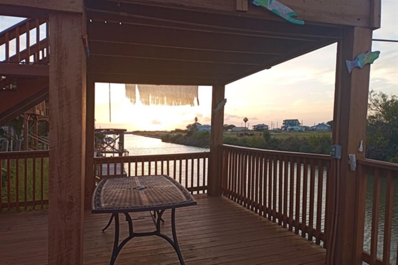 Covered wooden deck with a table overlooking a canal at sunset, with railing, nearby homes, and warm evening light.
