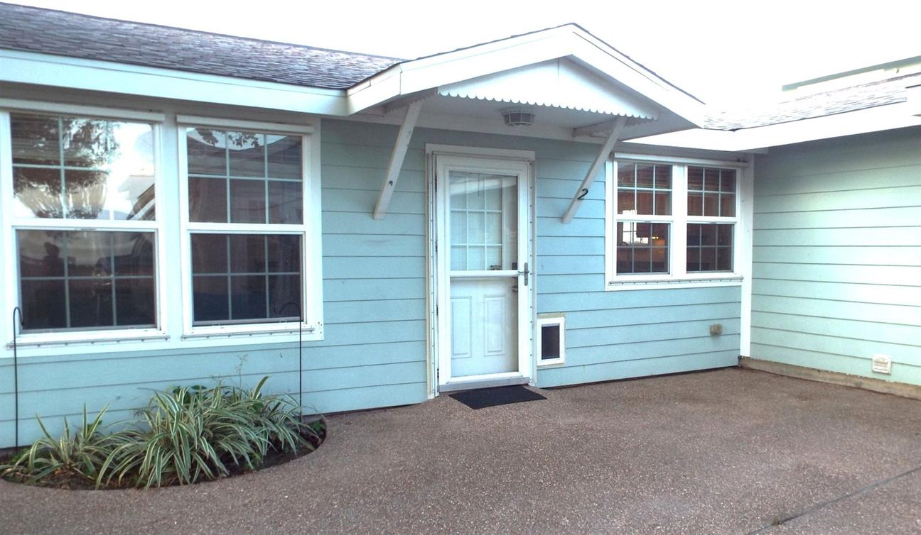 Light blue home exterior with white trim, front door under a small awning, windows on either side, and a concrete patio area.