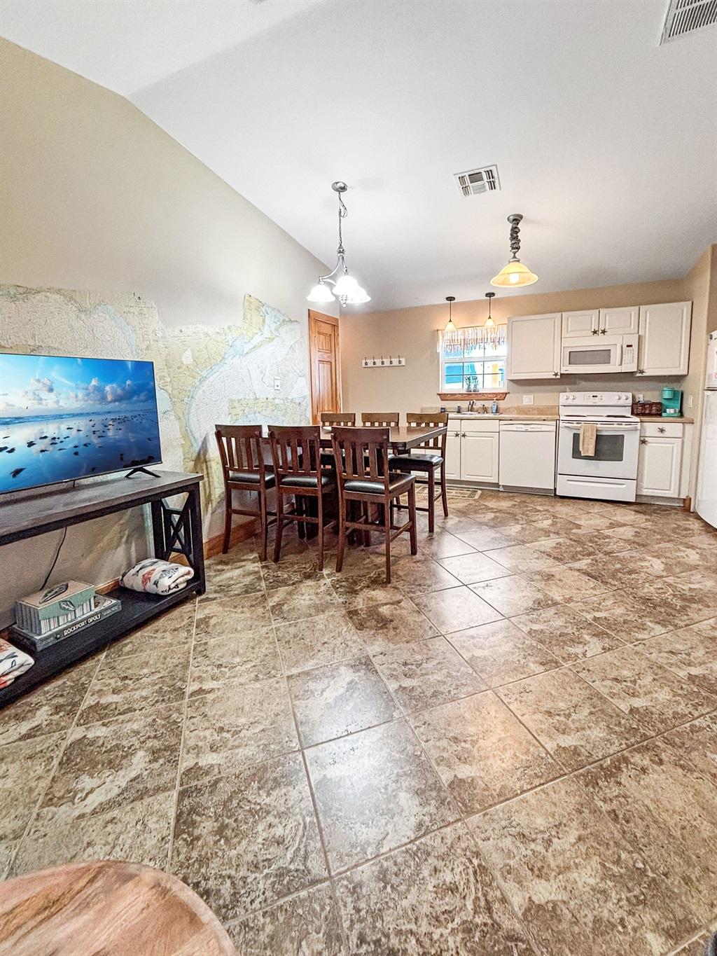 Wide view of a kitchen and dining space with a wooden table and chairs, white cabinets, appliances, wall art, and tile flooring.