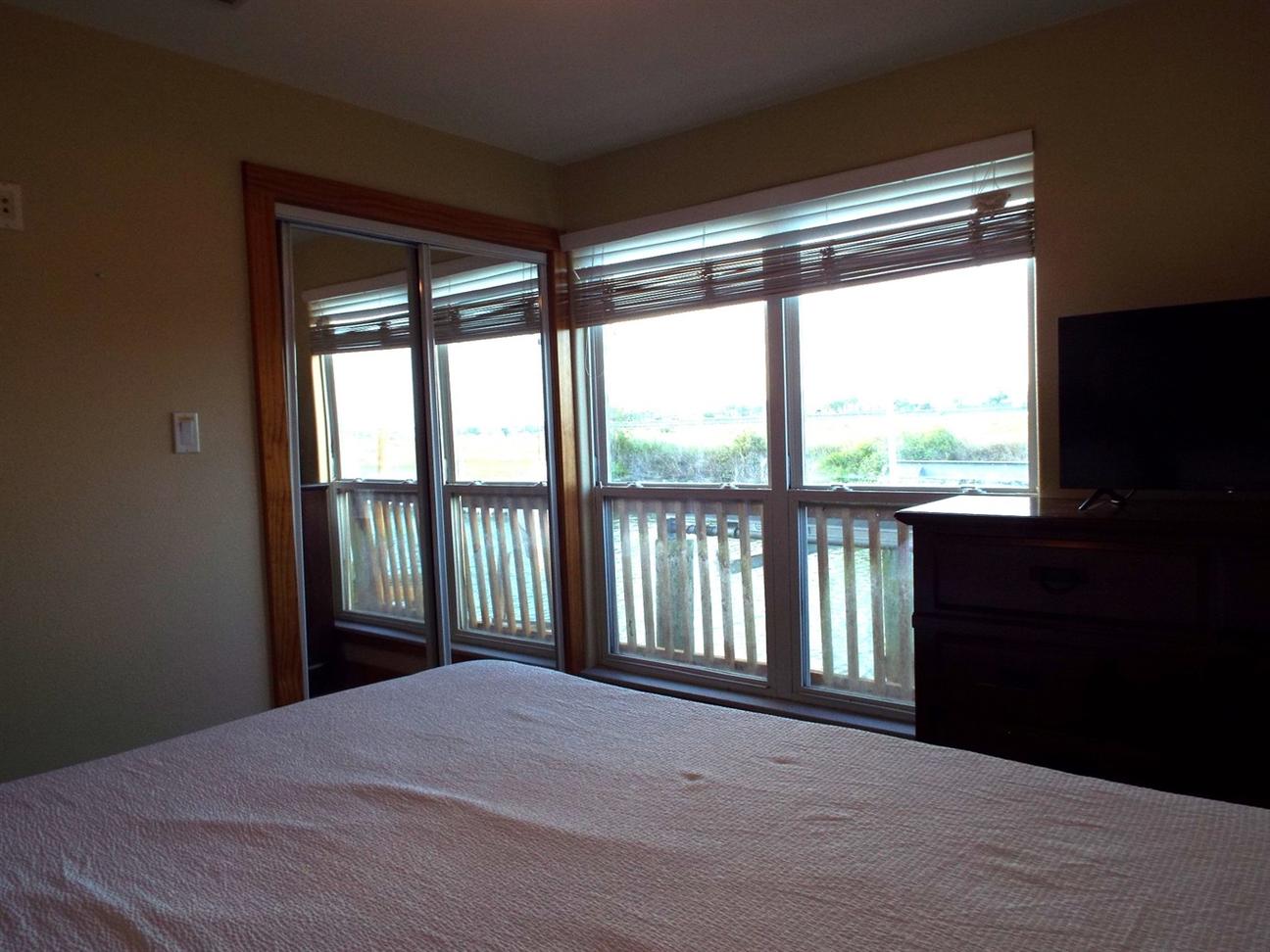 Bedroom with a bed facing large sliding glass doors and windows that open to a balcony overlooking the water, with a dresser and TV to the side.