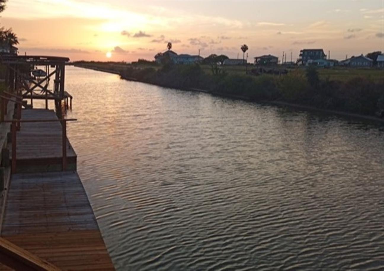 Sunset over a calm canal with wooden decks along the water and homes in the distance under a warm evening sky.