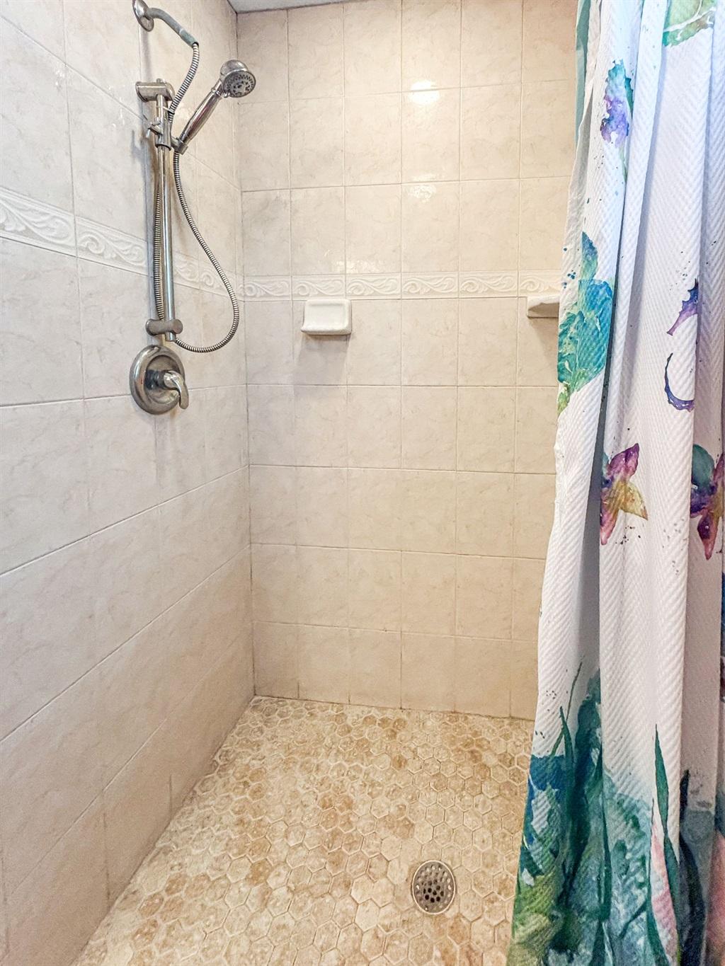 Tiled walk-in shower with handheld showerhead, built-in soap shelf, and pebble-style shower floor, partially covered by a patterned curtain.