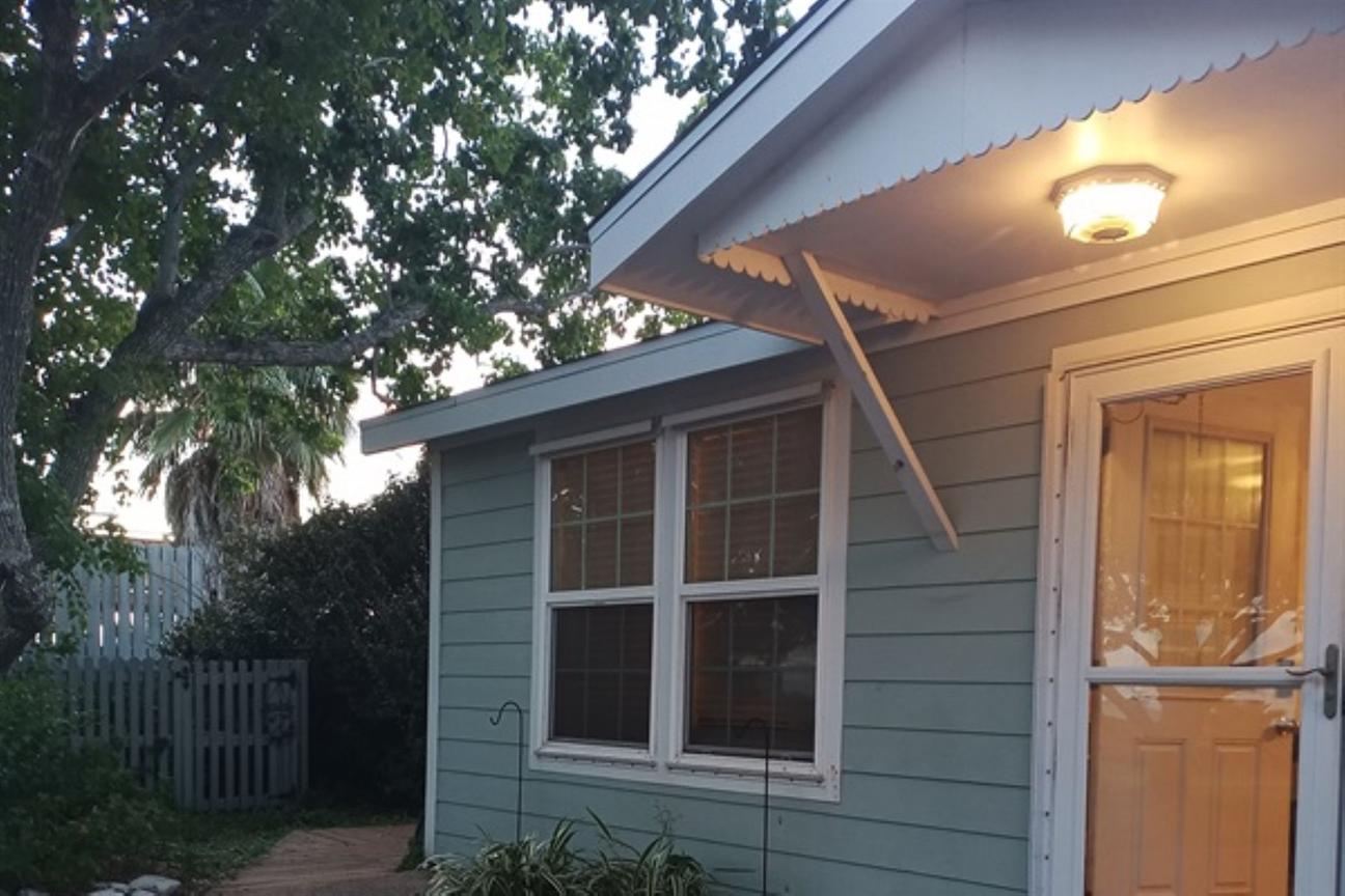 Light blue coastal home exterior with a lit porch light, window, and door, surrounded by trees and greenery at dusk.