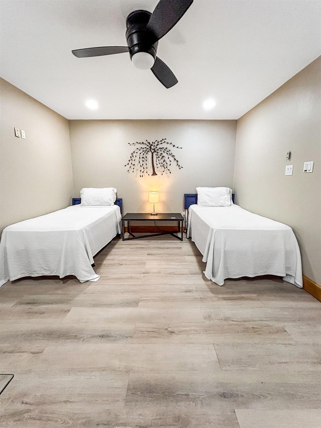 Bedroom with two twin beds, white bedding, a shared nightstand with a lamp, neutral walls, wood-look flooring, and a ceiling fan overhead.
