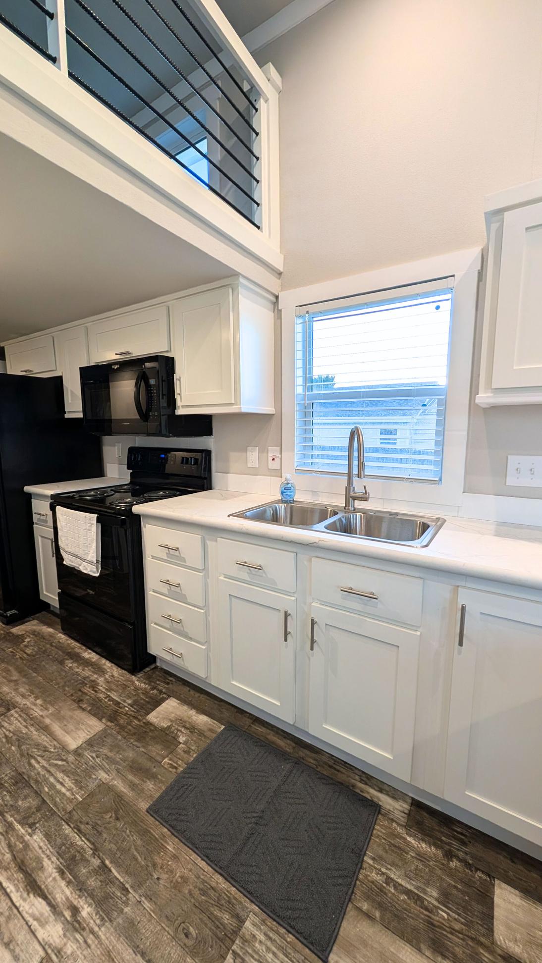 White kitchen cabinets with a double stainless steel sink under a window, black stove and microwave, and wood-look flooring.