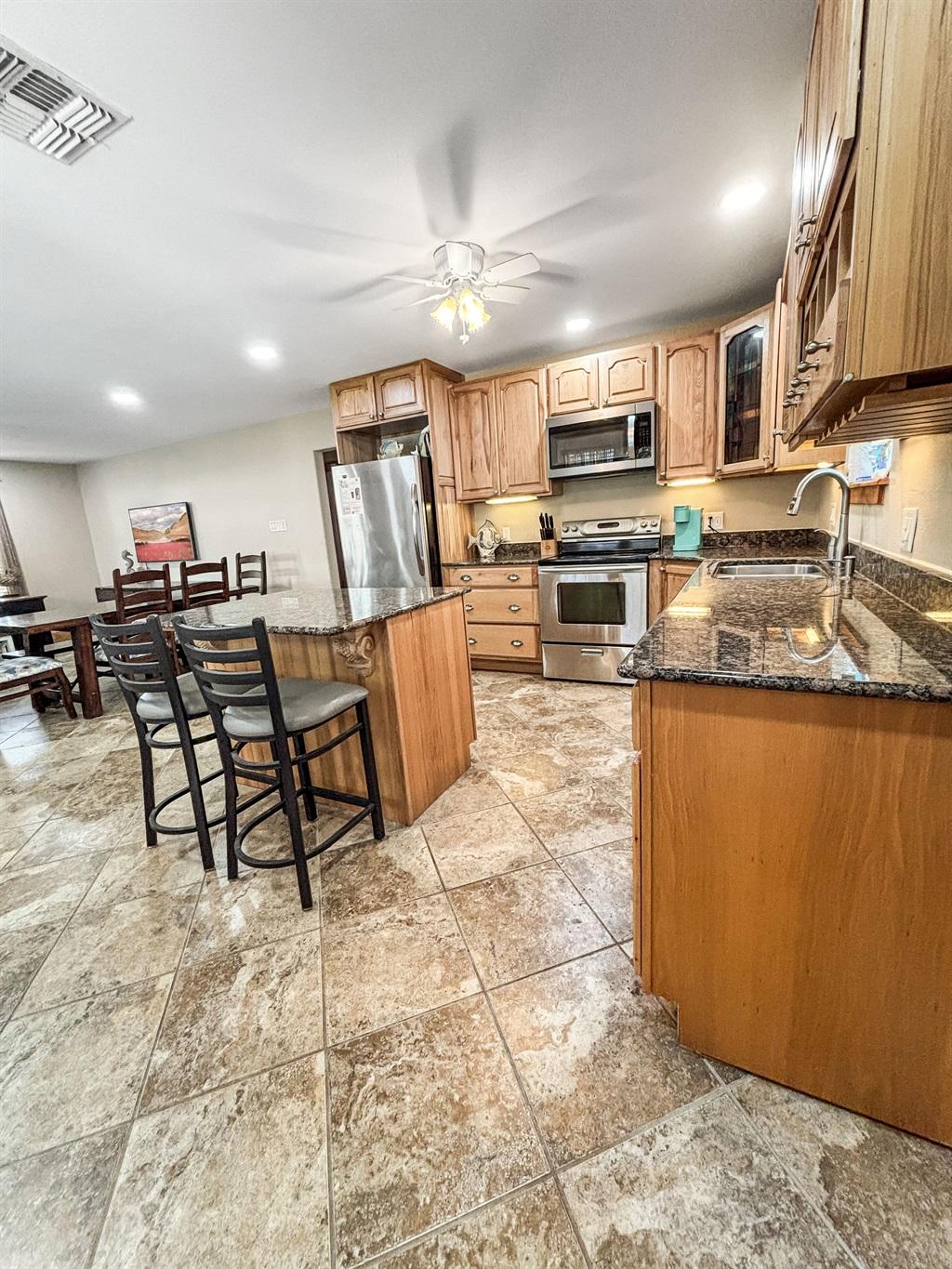 Spacious kitchen with wood cabinets, granite countertops, island with bar stools, stainless steel appliances, and tile flooring, open to a dining area.
