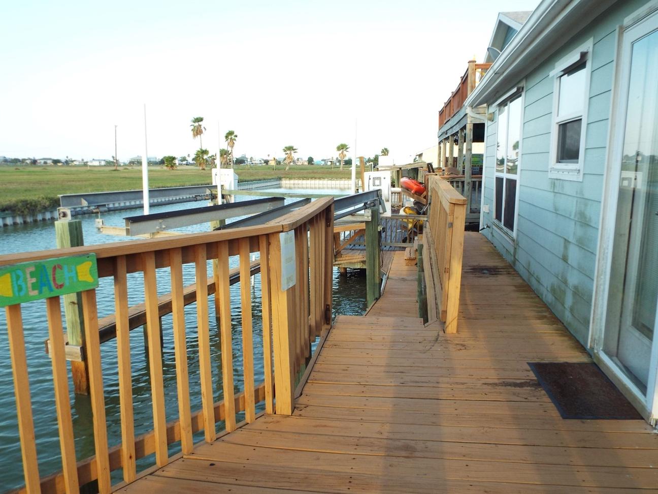Wooden deck along the water with railing, attached to a light blue coastal home, leading down to a dock with boats and canal views.