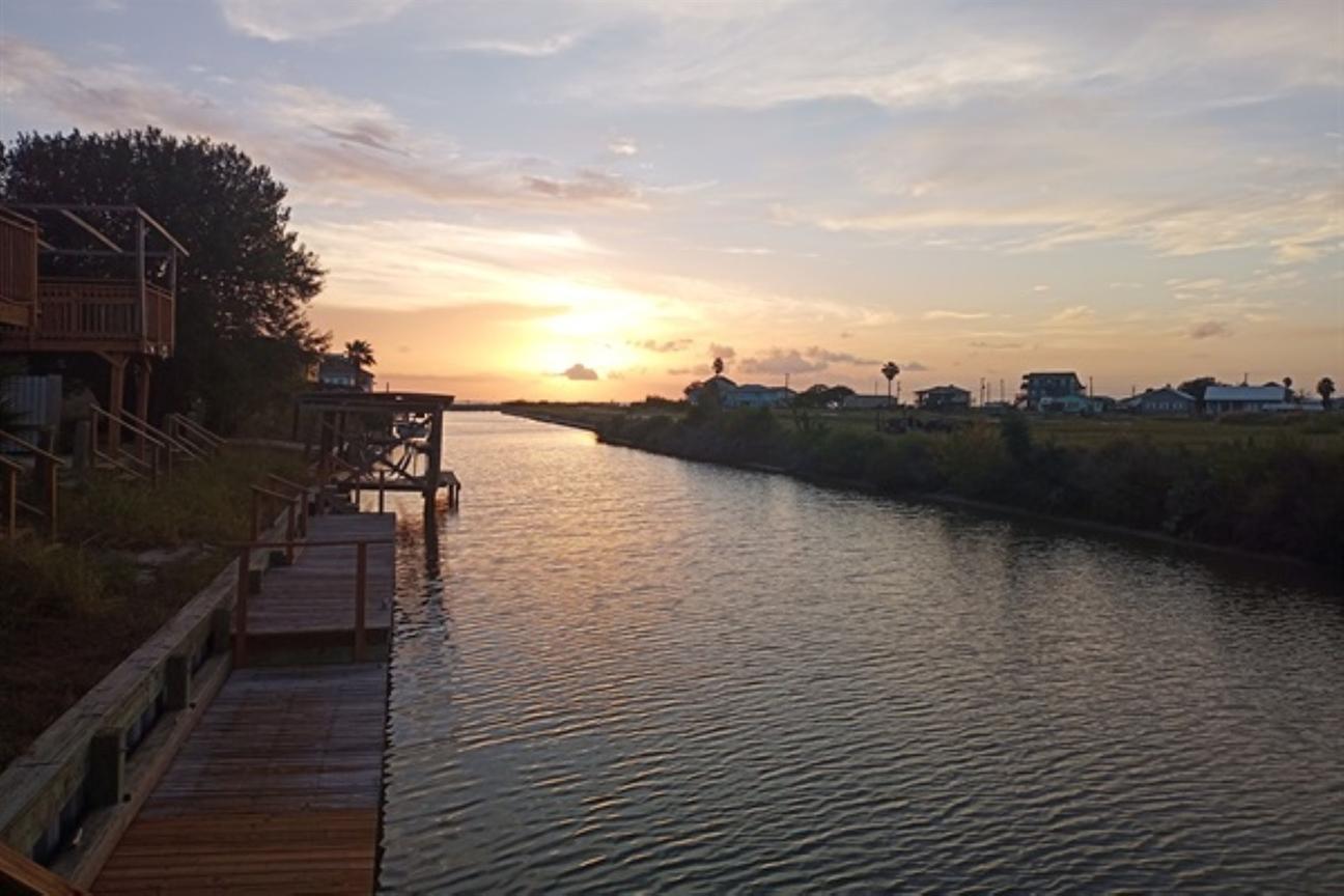 Sun setting over a calm canal with docks along the water and homes in the distance under a colorful evening sky.