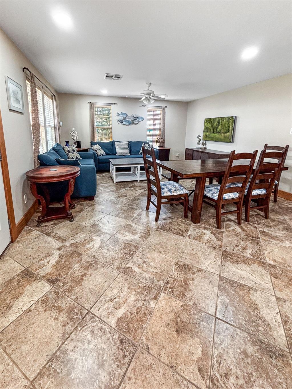 Large open room with tile flooring featuring a dining table with chairs, a blue sectional sofa, coffee table, and side table, with windows bringing in natural light.