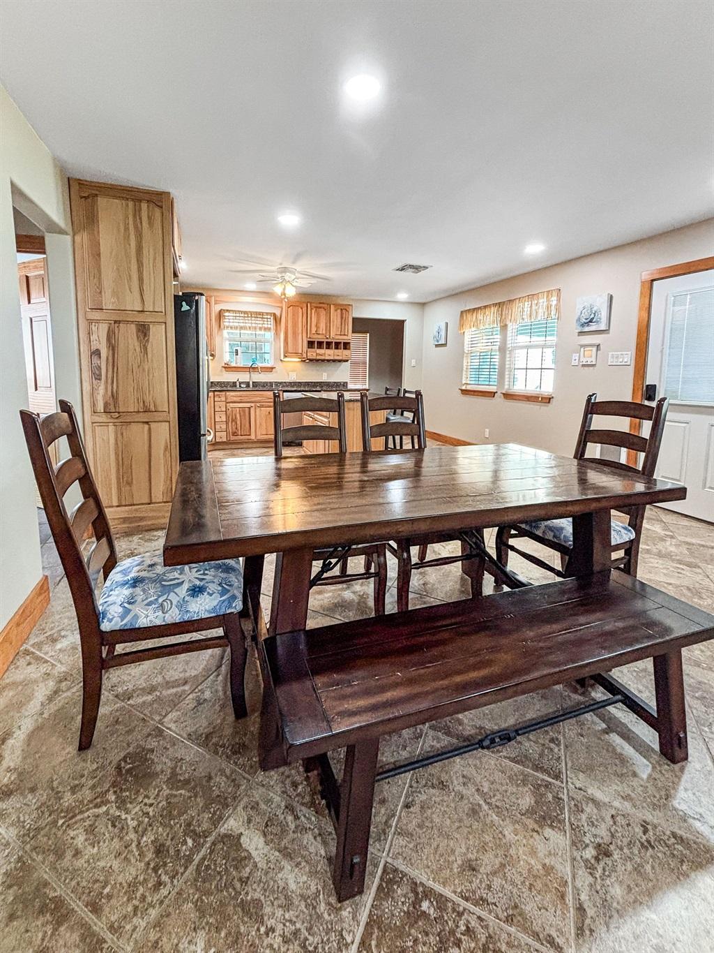 Dining area with a large wooden table, bench and chairs, tile flooring, and an open view into the kitchen with wood cabinetry.