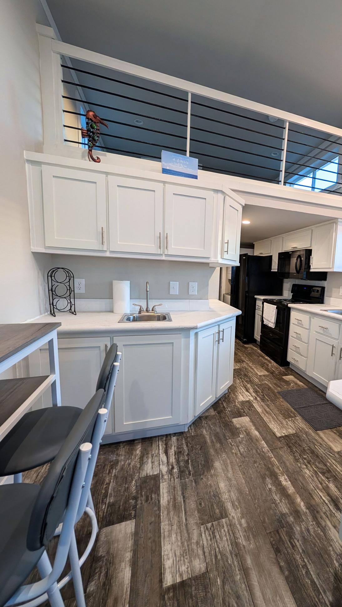 Modern kitchen with white cabinets, small sink area, breakfast bar seating, and a loft with railing above, leading into a galley kitchen with black appliances.