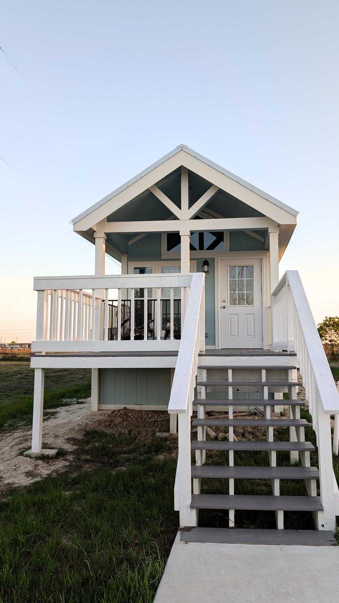 Front view of a raised coastal cottage with white railings, stairs leading up to a covered porch, and a light blue exterior under a clear sky.