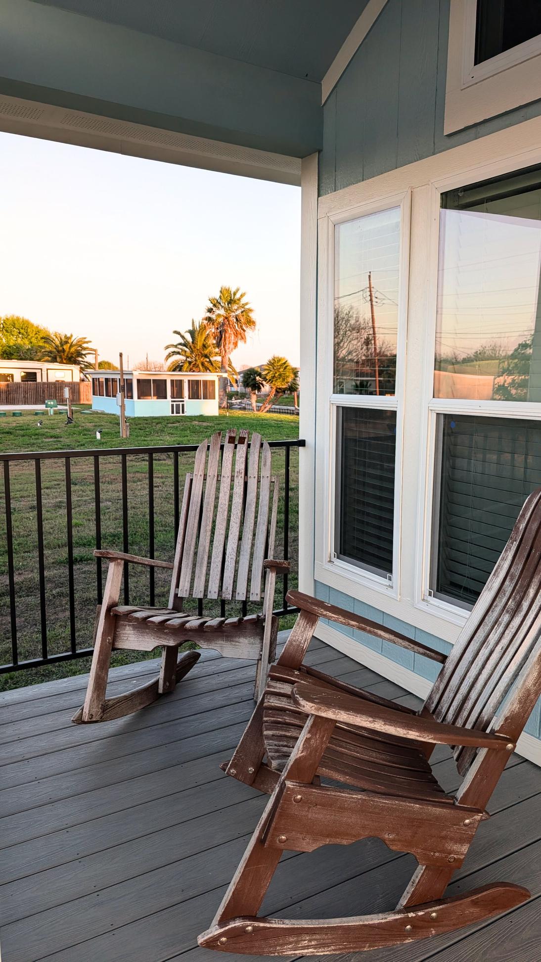 Two wooden rocking chairs on a covered porch overlooking a grassy yard with palm trees and coastal cottages in the background at sunset.