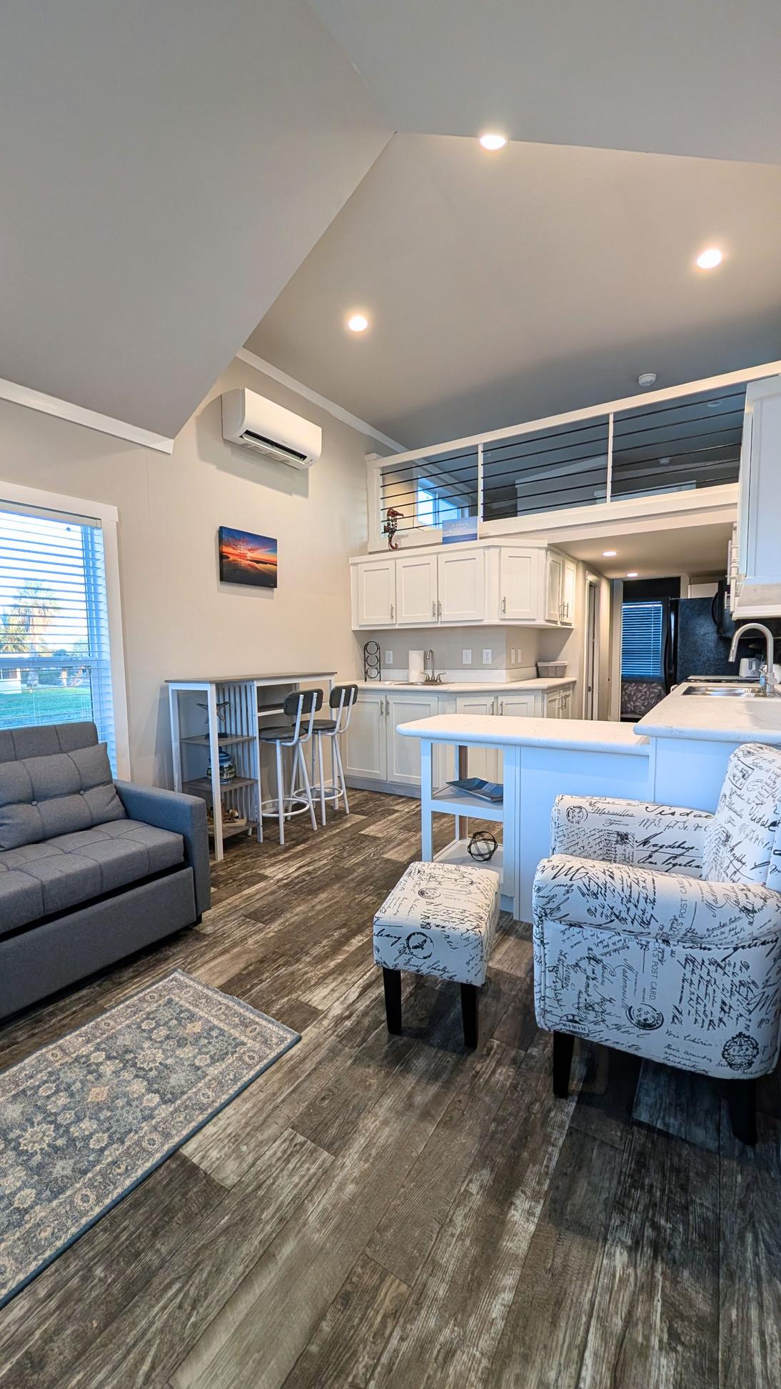 Open-concept living and kitchen area with a gray sofa, patterned chair and ottoman, breakfast bar with stools, white cabinetry, and a loft with railing above.