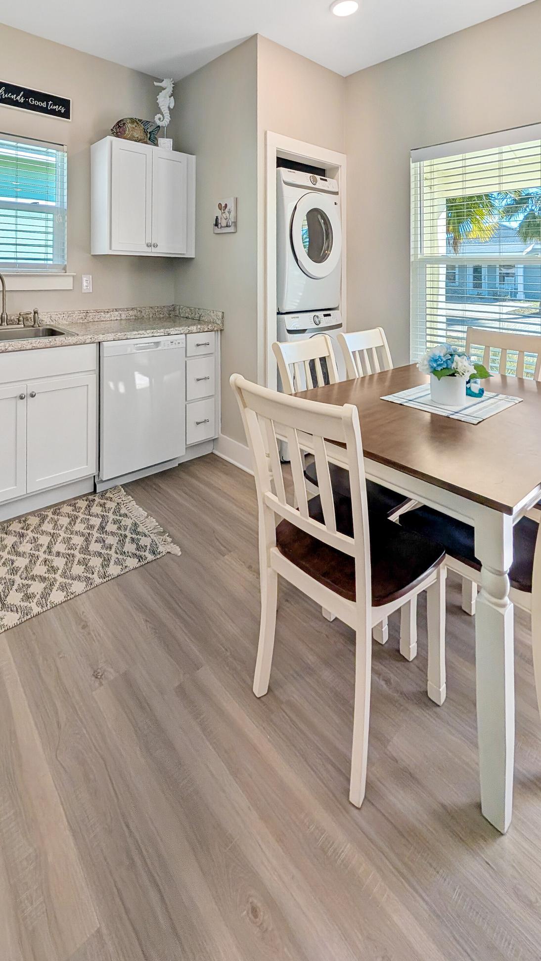 Dining area with a wooden table and white chairs next to a small kitchen with white cabinets, sink, dishwasher, stacked washer and dryer, and large windows with natural light.
