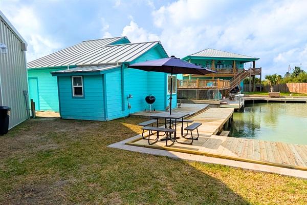 Canal-front backyard in Rockport, Texas with wooden dock, picnic table and umbrella, blue coastal home, and water access for boating and fishing.