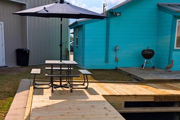 Wooden canal-front deck with picnic table and umbrella next to a bright blue coastal home, featuring a charcoal grill and outdoor space in Rockport, Texas.